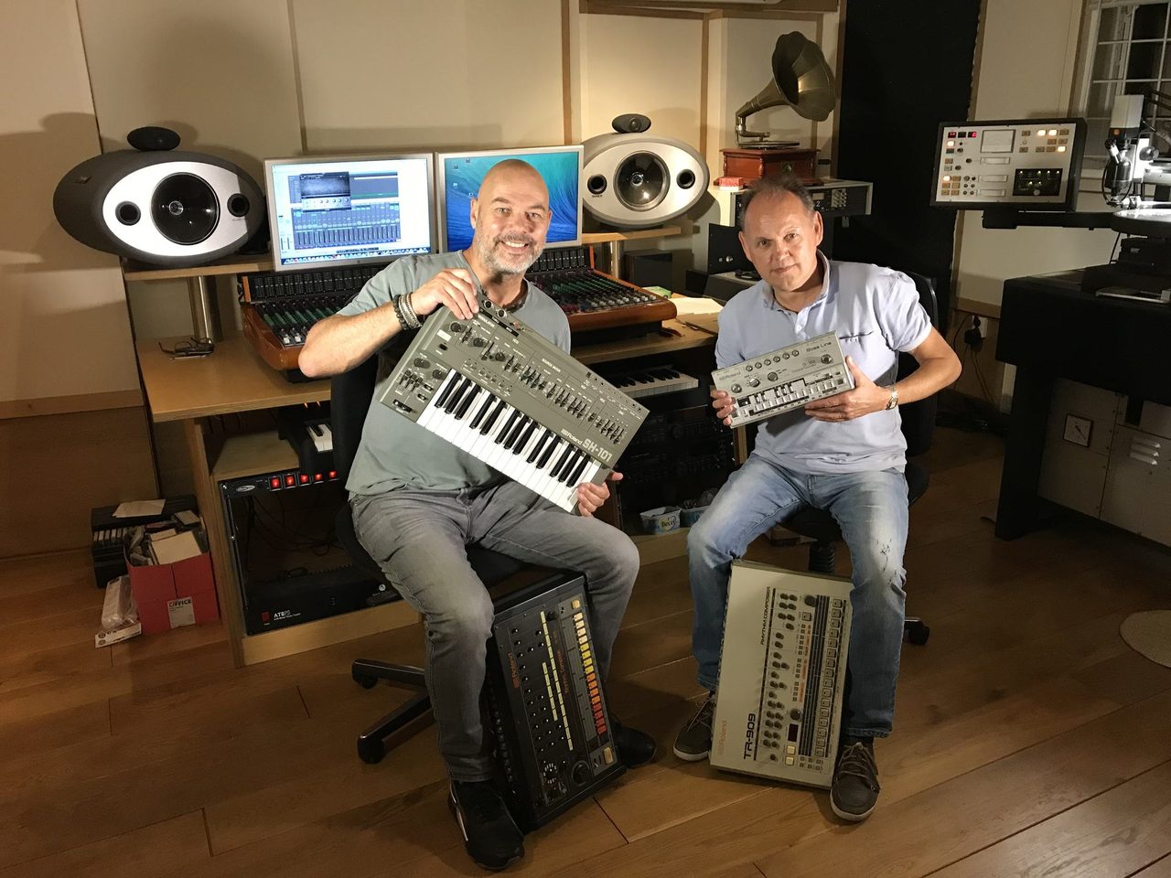 Lex van Coeverden and René van der Weijde in the studio, holding a Roland SH-101 and a TB-303, with a TR-808 and TR-909 at their feet.